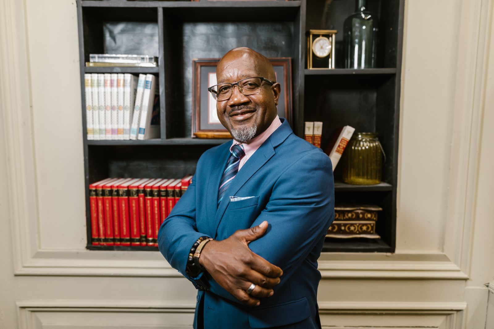 A confident, smiling older businessman in a blue suit standing with arms relaxed in front of a bookshelf, looking warm and self-assured.