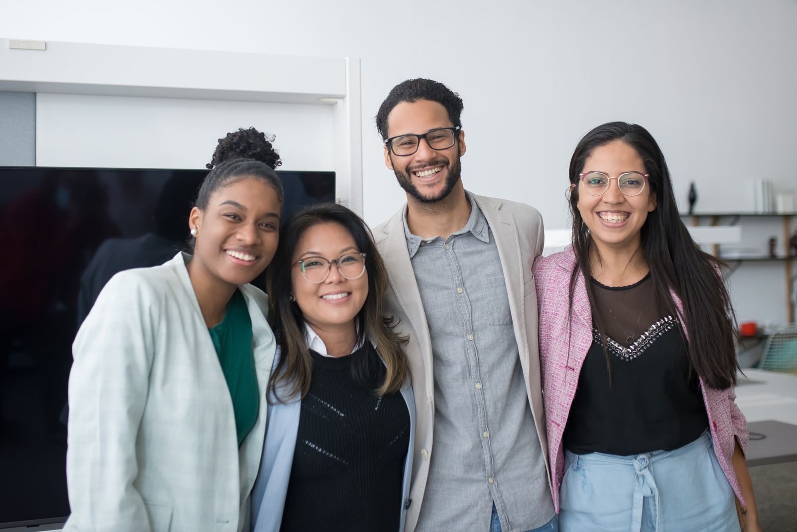 Four smiling coworkers standing close together with arms around each other in a bright office.